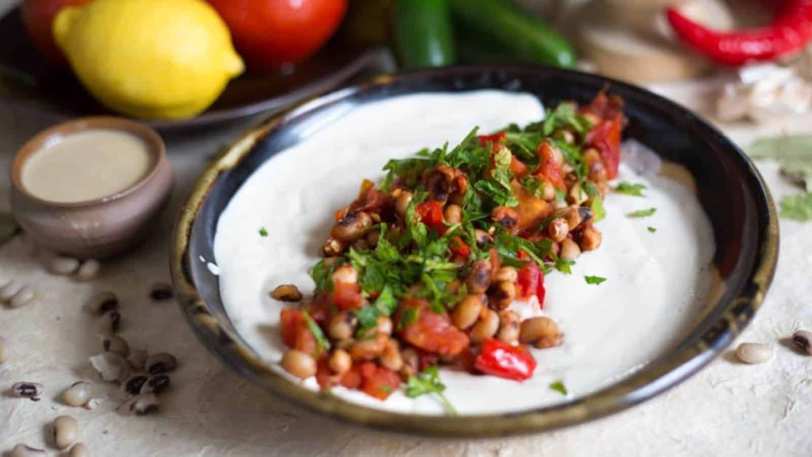 A plate of creamy hummus topped with a mix of black-eyed peas, diced tomatoes, red peppers, and parsley. Surrounding the dish are whole lemon, tomato, cucumber, red pepper, and a small bowl of sauce.