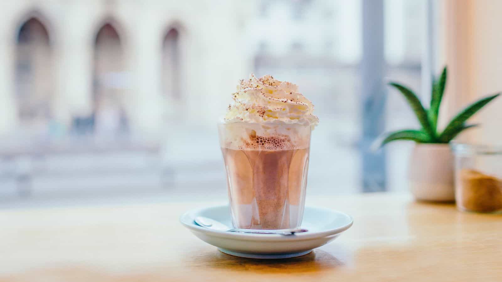 A glass of hot chocolate topped with whipped cream and sprinkles sits on a white saucer with a spoon. The background is softly blurred, showing part of a window and a small potted plant on a wooden table.