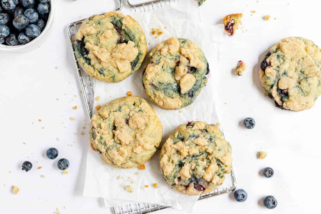 Four blueberry cookies rest enticingly on a cooling rack lined with parchment paper, surrounded by loose blueberries and crumbs. A bowl of blueberries teases from the top left corner, promising that these good cookies will disappear in minutes.