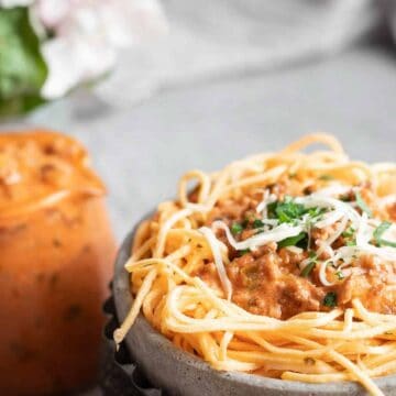 A bowl of spaghetti topped with meat sauce and garnished with shredded cheese and herbs. In the background, a jug filled with additional meat sauce is visible. A blurred floral arrangement is partially visible to the left.