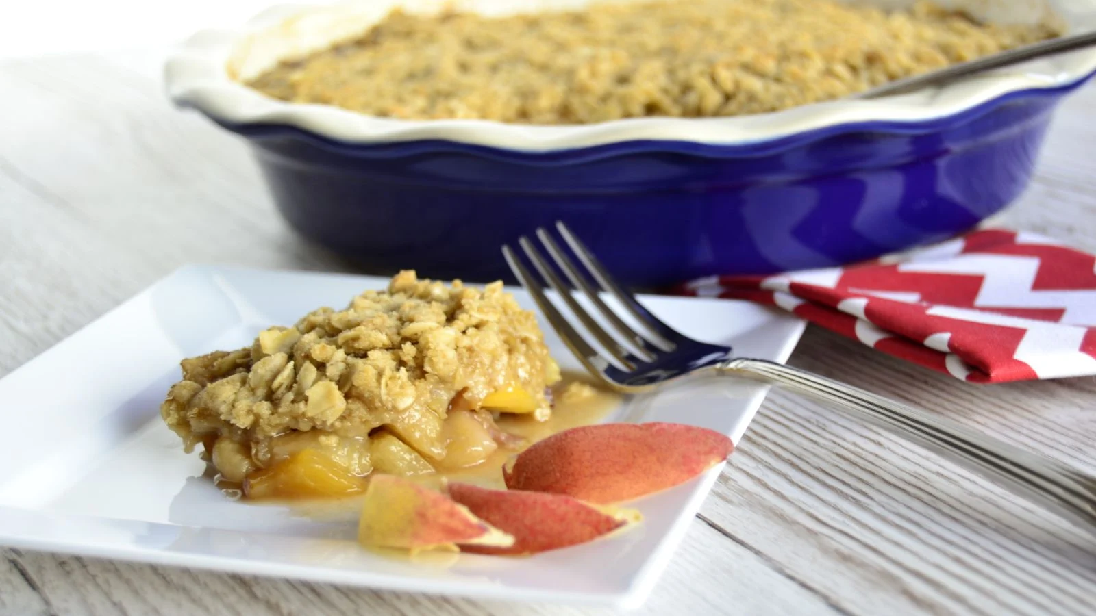 A slice of fruit crumble, a true dreamy dessert, sits on a white plate with two peach slices and a fork beside a blue baking dish filled with the same treat. In the background, a red and white chevron napkin rests on the wooden surface, ready to wow any crowd.