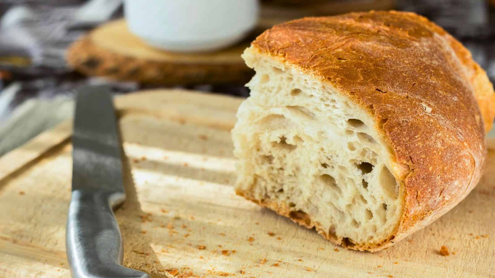 A close-up of a half-loaf of rustic bread on a wooden cutting board. A serrated knife with a silver handle is placed beside the bread. The bread has a crusty exterior and a soft, airy interior. A blurred background shows more kitchen items.