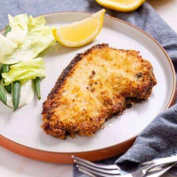 A plate with a breaded pork chop, a wedge of lemon, and a side of lettuce and green beans. A blue cloth napkin and utensils are beside the plate.