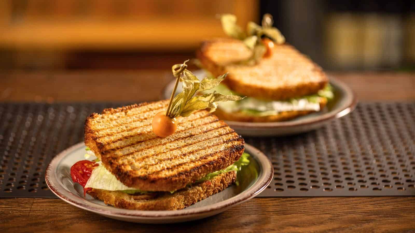 Two grilled sandwiches are on separate plates. Each sandwich is filled with lettuce, tomato, and other ingredients, and topped with a physalis. The sandwiches are placed on a wooden surface with a blurred background.