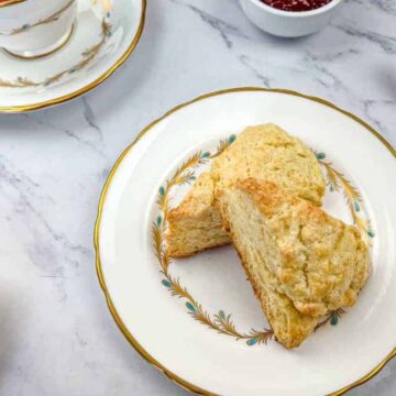 A plate with two triangular scones on a marble surface. The plate features a decorative pattern. Beside it, there is a butter knife and a small bowl of jam. A cup on a saucer is partially visible to the left.