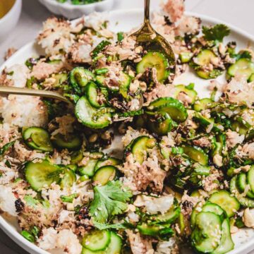 A close-up of our Crispy Cucumber Rice Salad Recipe features sliced cucumbers mixed with rice, herbs, and spices, garnished with cilantro on a white plate. A spoon is scooping a portion, revealing additional garnishes in the background.