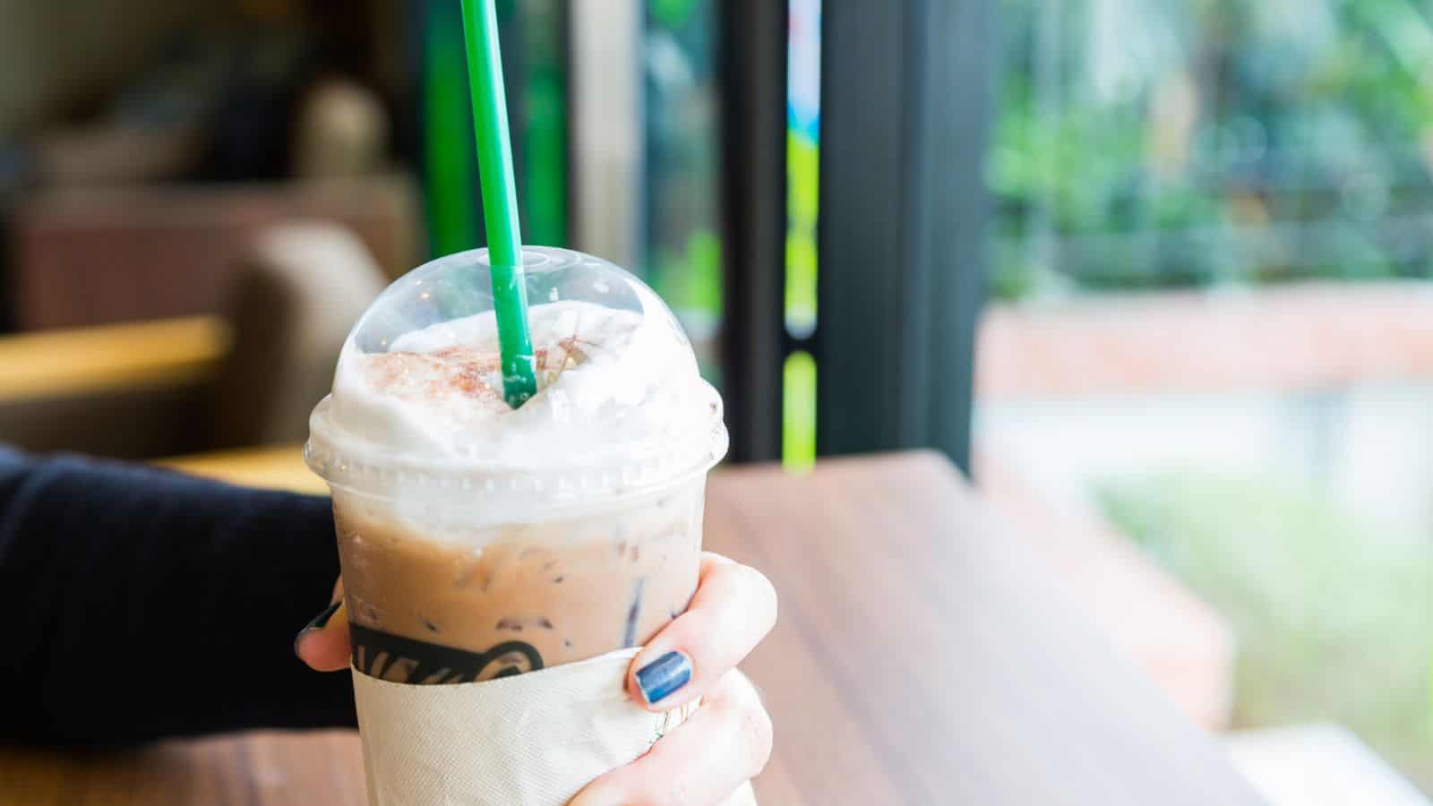 A hand with painted nails holds an iced coffee drink topped with whipped cream and a green straw. The drink is in a plastic cup with a sleeve on it. The background shows a blurred indoor setting with a window and greenery outside.