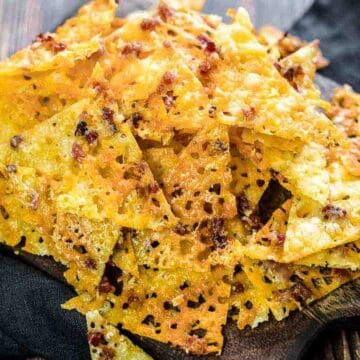 A pile of crispy cheese chips with a golden-brown color is placed on a wooden board with a dark cloth underneath. The chips have a textured appearance, showing small holes and bits of melted cheese.