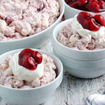 Bowls of cherry and marshmallow dessert salad are arranged on a table with a red and white checkered cloth and metal spoons. The dessert is topped with whipped cream and cherries.