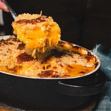 A spoon lifts a portion of baked macaroni and cheese with a crispy breadcrumb topping from an oval black casserole dish. The dish rests on a wooden trivet, and a blue cloth is partially visible in the background.