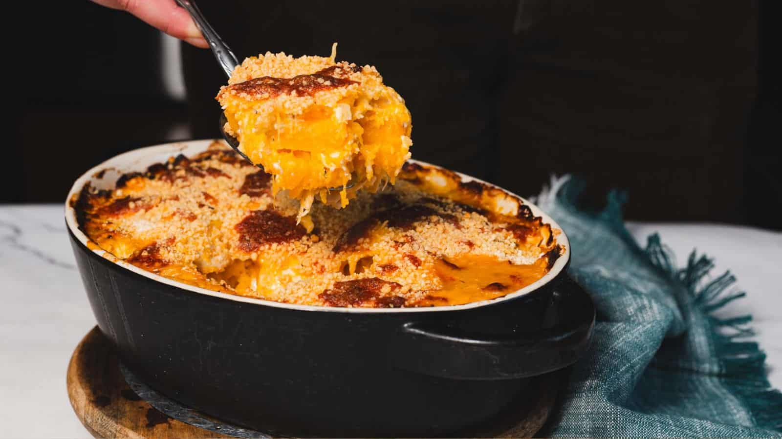 A person lifts a spoonful of cooked macaroni and cheese from an oval black baking dish. The dish is topped with a breadcrumb crust, and a blue cloth is partially visible underneath. The table surface is white.