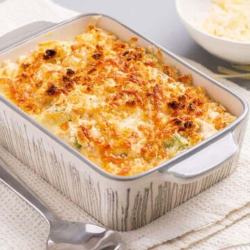 A baked dish with a melted golden-brown cheese crust is in a rectangular ceramic baking dish. It rests on a textured towel, with a spoon nearby. A small bowl of shredded cheese is positioned in the background.