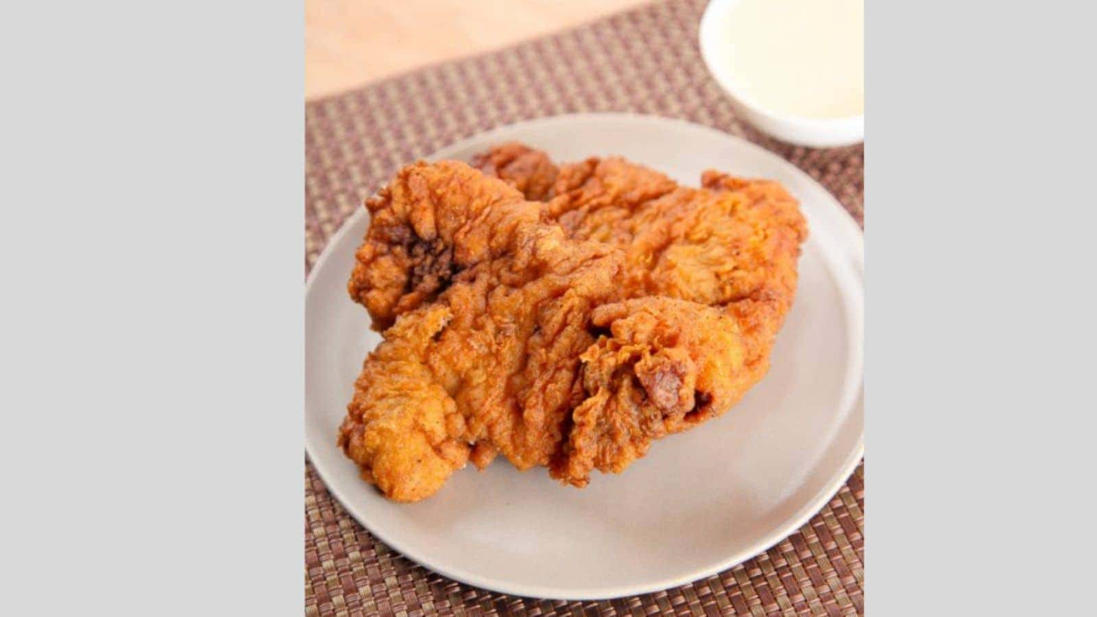 A plate with a piece of crispy fried chicken sits on a woven placemat, evoking sweet memories of Grandma's recipes. A small bowl of creamy white sauce lingers in the background.