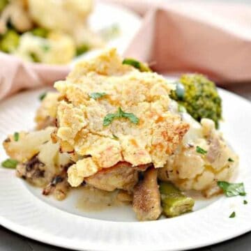 A dish featuring baked chicken with a crispy topping, embodying cozy meals, is served on a white plate. Accompanied by broccoli and cauliflower, the plate rests beside a pink cloth napkin on a gray surface. Another plate is visible in the background, adding to the inviting setting.