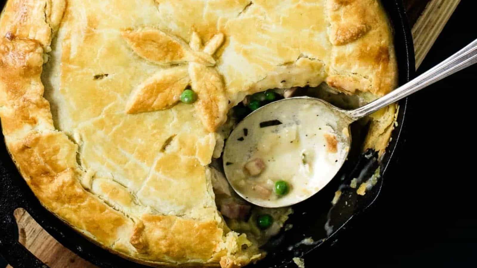 A golden-brown chicken pot pie with a section removed, revealing a creamy filling with peas and diced meat. A silver spoon rests in the empty part of the pie, and leaf decorations adorn the crust.