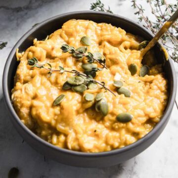 A gray bowl holds a creamy, golden chicken pumpkin risotto topped with crunchy pumpkin seeds and fresh thyme sprigs. A spoon rests invitingly in the bowl, all set on a cool marble surface.