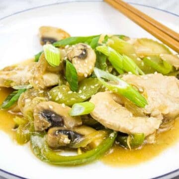 A plate of stir-fried chicken with sliced mushrooms, bell peppers, and green onions, topped with sesame seeds. A pair of wooden chopsticks rests on the edge of the white plate with a dark rim, placed on a marble surface.