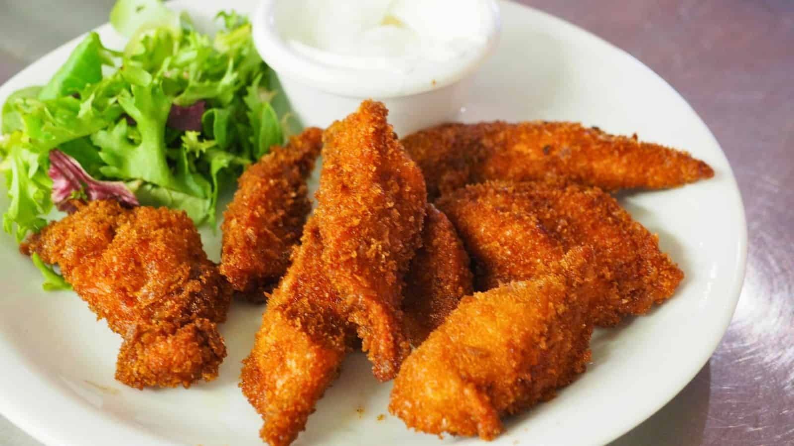 A plate of golden-brown breaded chicken strips is served with a side of mixed greens and a small container of white dipping sauce.