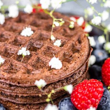 A stack of chocolate waffles on a plate is surrounded by fresh raspberries, blueberries, and small white flowers for decoration, creating a dreamy dessert that's sure to please any crowd.