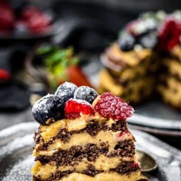 A slice of Heavenly Cakes' layered chocolate and cream cake, perfectly crafted to impress, is topped with blueberries and raspberries on a plate. In the background, strawberries blur softly. A dusting of powdered sugar adds the final touch for any sweet tooth craving delight.