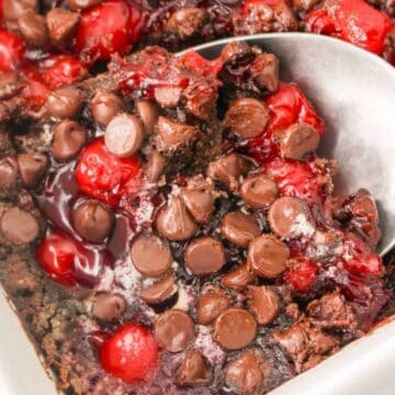 A close-up of a dessert being served from a baking dish showcases rich layers of chocolate chips and red cherry filling. This irresistible chocolate treat tempts with every spoonful lifted, making you dream of licking the bowl clean.