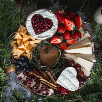 A charcuterie board with an assortment of cheeses, including heart-shaped brie with pomegranate seeds, sliced cheddar, and white cheese. It features strawberries, blueberries, grapes, and crackers alongside the rich texture of crème brûlée and is surrounded by fragrant pine greenery.