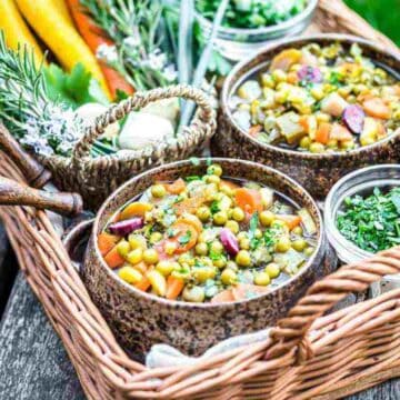 A wicker basket on a wooden table showcases two bowls of vegetable stew, a remarkable dish in the world of recipes. The hearty stew, filled with peas, carrots, and assorted vegetables, exudes the wow factor. Beside the bowls are smaller containers with chopped herbs, fresh carrots, and rosemary.