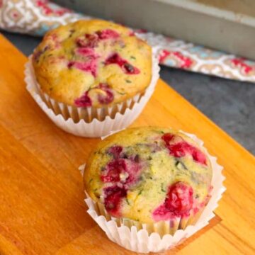 Two muffins with visible berries are placed in paper liners on a wooden cutting board. A baking tray with another muffin is partially visible in the background, along with a patterned cloth. The surface is dark and smooth.