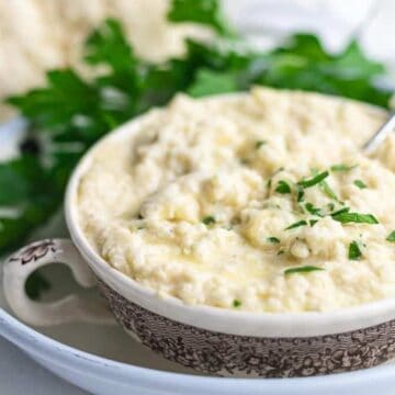 A bowl of creamy mashed cauliflower, an unexpected twist on classic side dishes, is garnished with chopped parsley and sits on a decorative plate. A spoon rests inside, ready to serve this guaranteed hit, with fresh parsley artfully placed beside it.