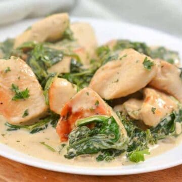 A white plate with creamy chicken breast pieces, spinach, and herbs, served in a light cream sauce. The dish is placed on a wooden surface with a soft green cloth partially visible in the background.