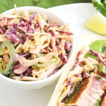 A close-up of a plate showcases a bowl of coleslaw with jalapeños alongside a taco filled with sliced meat and cilantro. A lime wedge and cilantro garnish pair beautifully, all set against a green napkin background, making it the perfect comfort food duo.