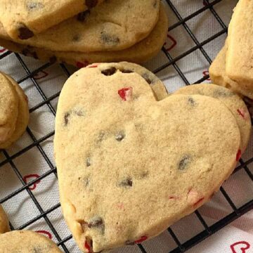 Heart-shaped cookies with chocolate chips and red bits are on a black cooling rack, resting on a cloth with small red hearts. A partial view of a pink rose is visible in the corner.