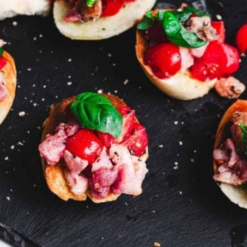 Slices of bread topped with diced tomatoes, chopped meat, and fresh basil are arranged on a black slate surface.