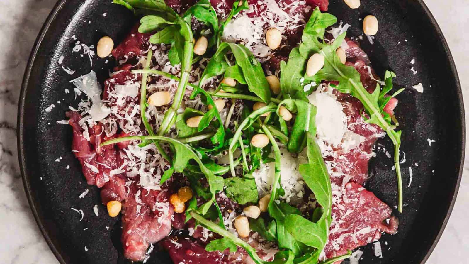 A black plate features a serving of beef carpaccio topped with arugula, shaved Parmesan cheese, pine nuts, and drizzled with olive oil, set on a marble surface.