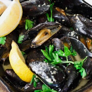 A black skillet contains cooked mussels in a creamy sauce, garnished with fresh parsley. Two lemon wedges are placed on top. A spoon rests in the dish. Pieces of bread are visible on the side, partially out of view.