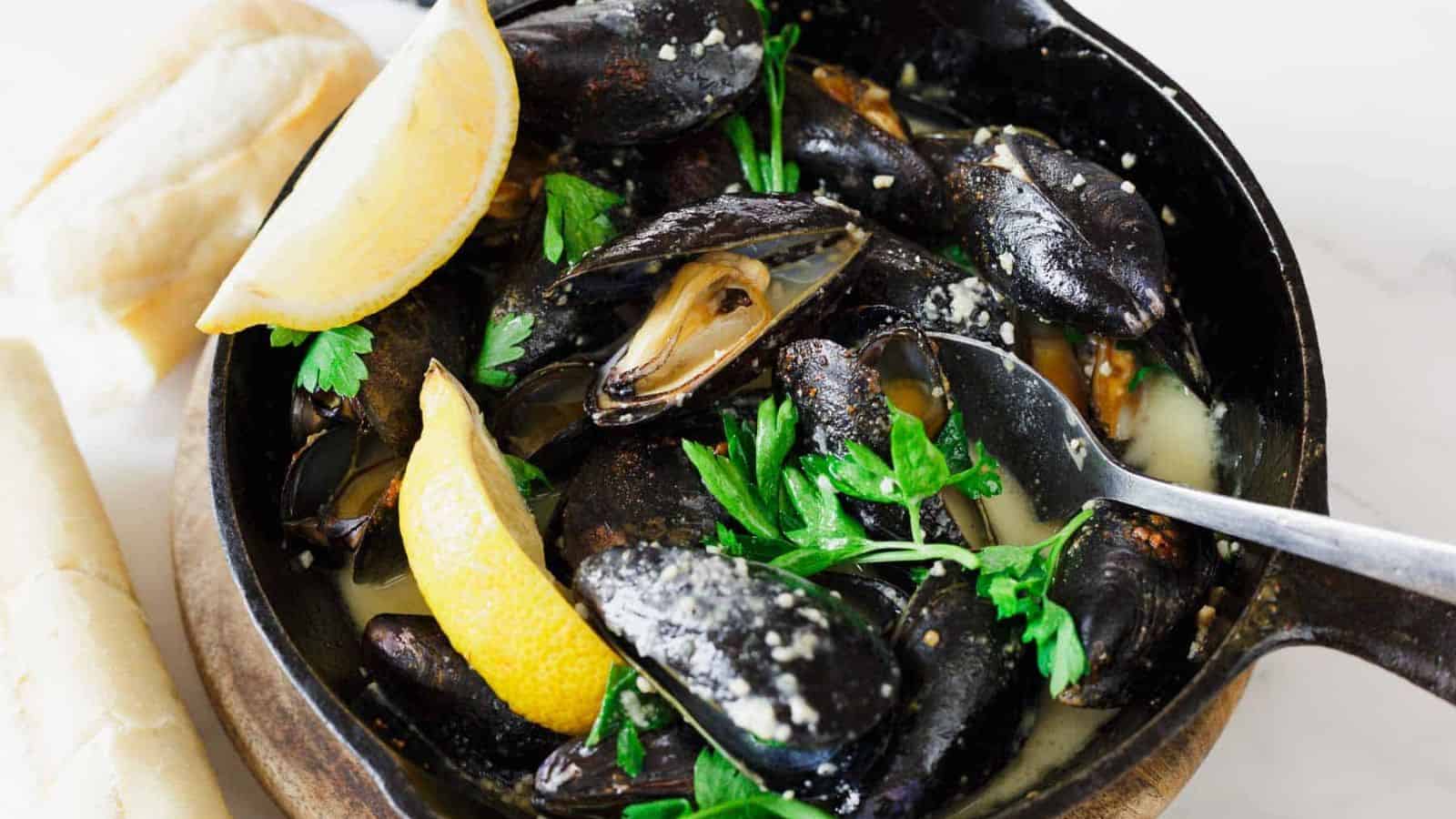 A skillet contains cooked mussels in a light sauce, garnished with parsley. A lemon wedge is placed inside, and a spoon rests in the dish. Partially visible bread rolls are on the side.