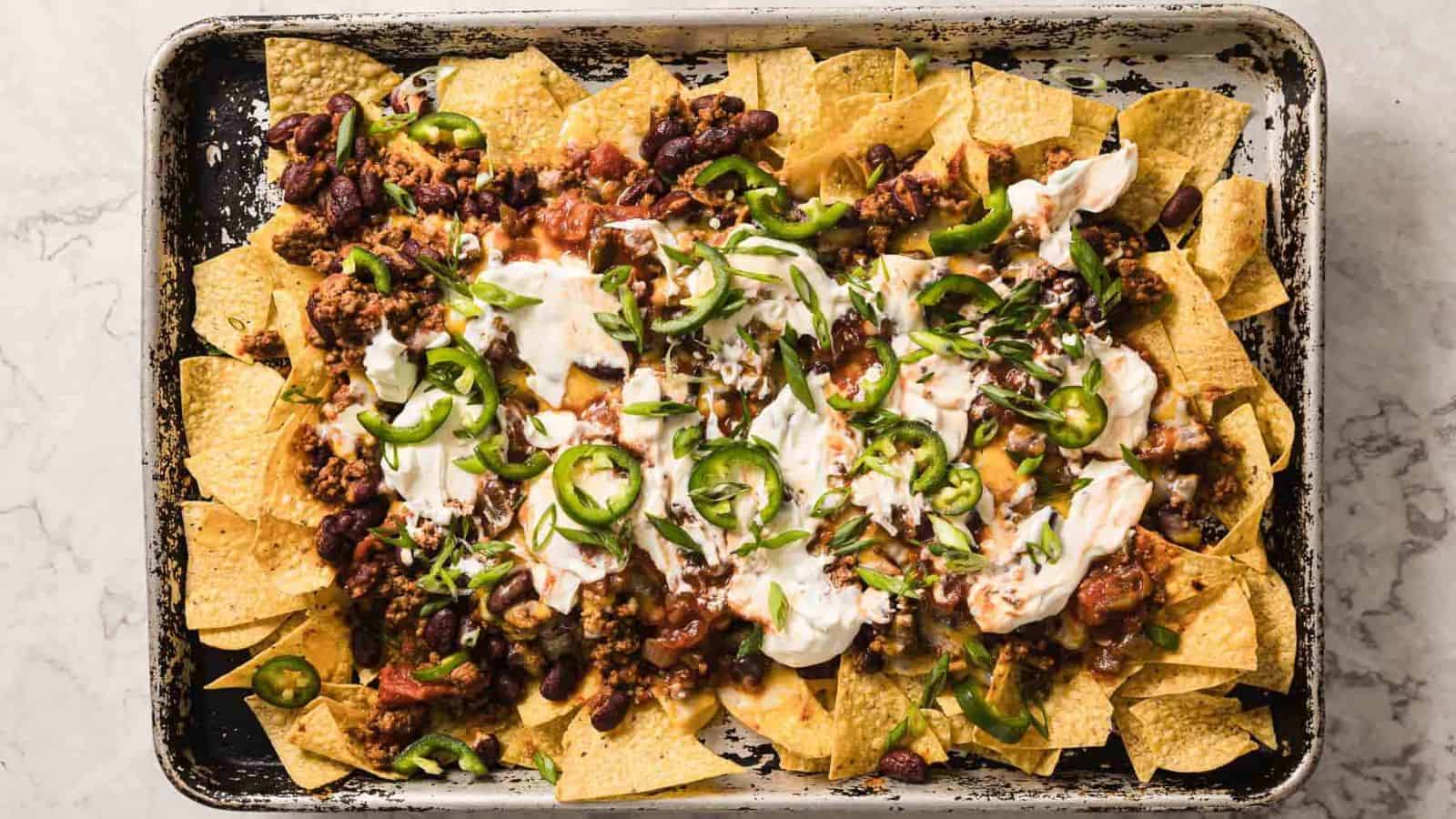 A baking sheet filled with nachos topped with melted cheese, ground meat, beans, jalapeño slices, sour cream, and chopped green onions. The nachos are arranged in an overlapping pattern with ingredients spread evenly.