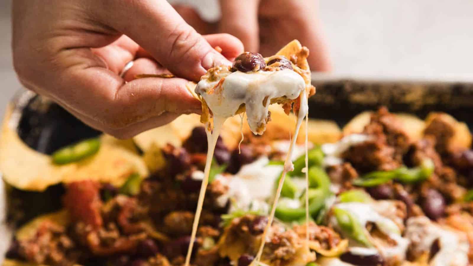 A close-up of a hand holding a tortilla chip topped with melted cheese, meat, beans, and green onions. The chip is pulled from a tray of loaded nachos, with strings of cheese stretching from the tray.