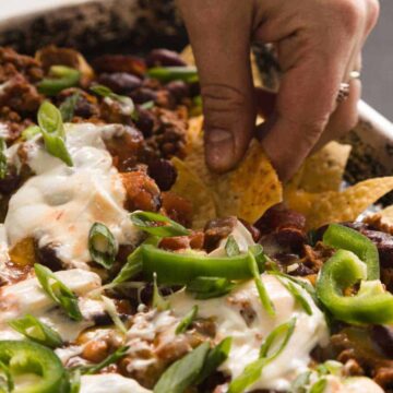 A hand is reaching for a chip from a tray of loaded nachos. The nachos are topped with melted cheese, ground meat, sliced jalapeños, sour cream, and green onions. The dish sits on a dark baking sheet.