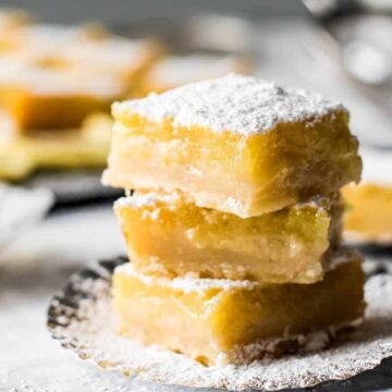 A stack of three lemon bars dusted with powdered sugar is placed on a metal plate. In the background, more lemon bars are visible on a blurred tray and scattered sugar is sprinkled around the scene.