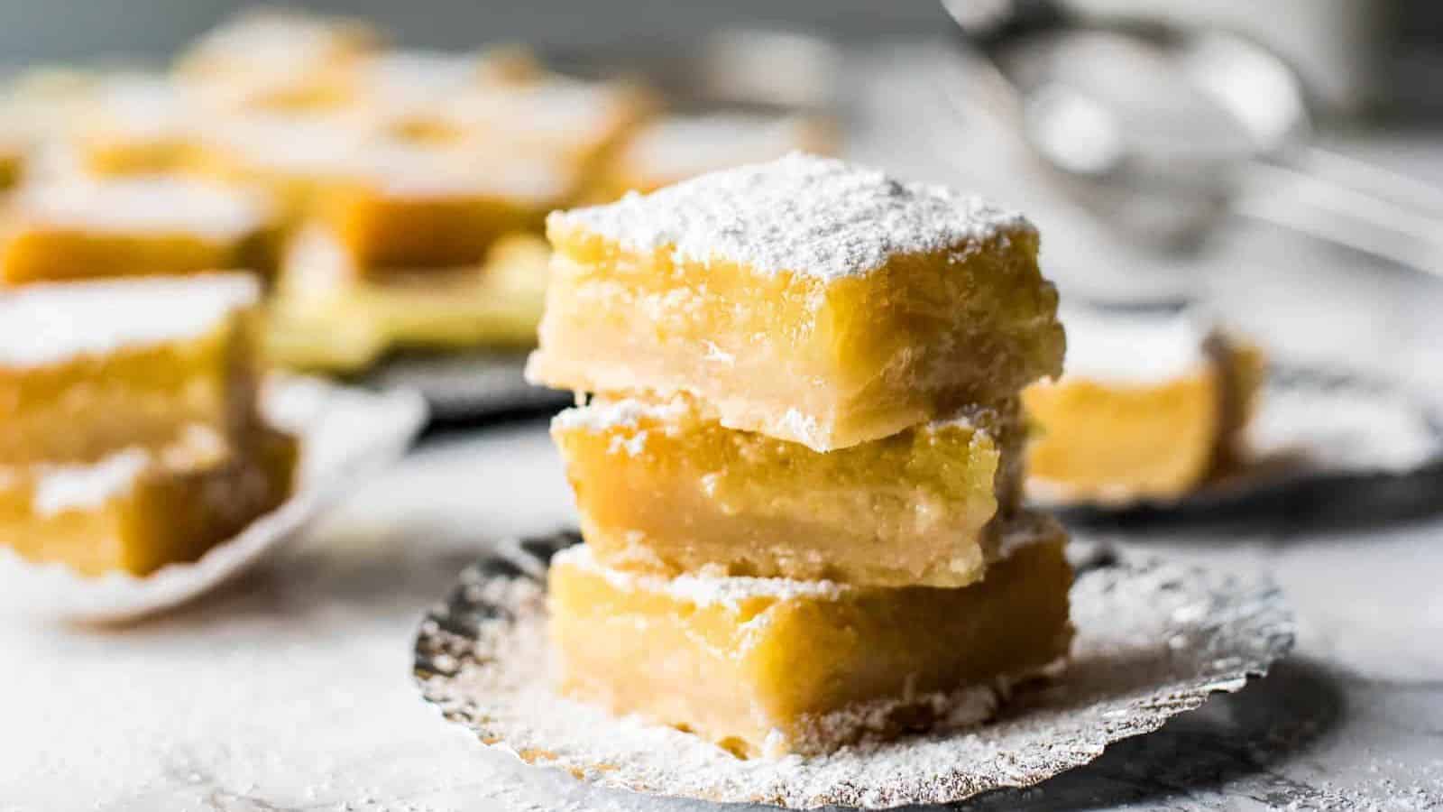 A stack of three lemon bars on a plate with more lemon bars in the background.