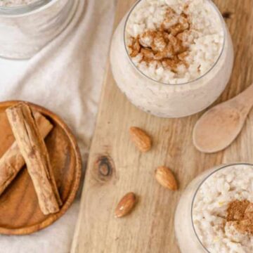 Two glasses filled with rice pudding topped with cinnamon sit on a wooden board. A wooden spoon is placed nearby, alongside a small plate with cinnamon sticks and almonds. A jar of rice lingers in the background, evoking the charm of classic recipes that remind us of forgotten flavors.