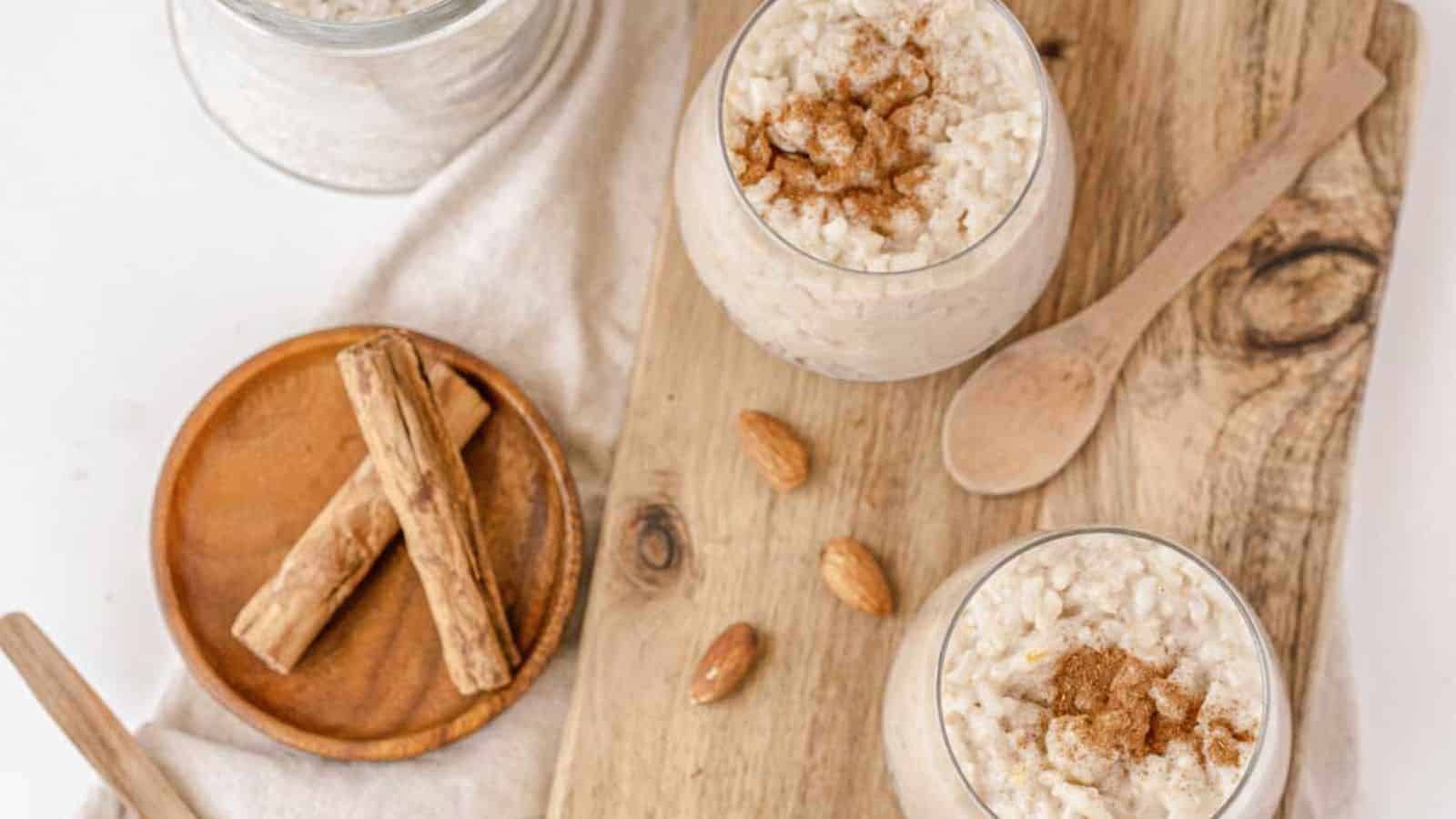 Two glasses filled with rice pudding topped with cinnamon sit on a wooden board. A wooden spoon is placed nearby, alongside a small plate with cinnamon sticks and almonds. A jar of rice lingers in the background, evoking the charm of classic recipes that remind us of forgotten flavors.