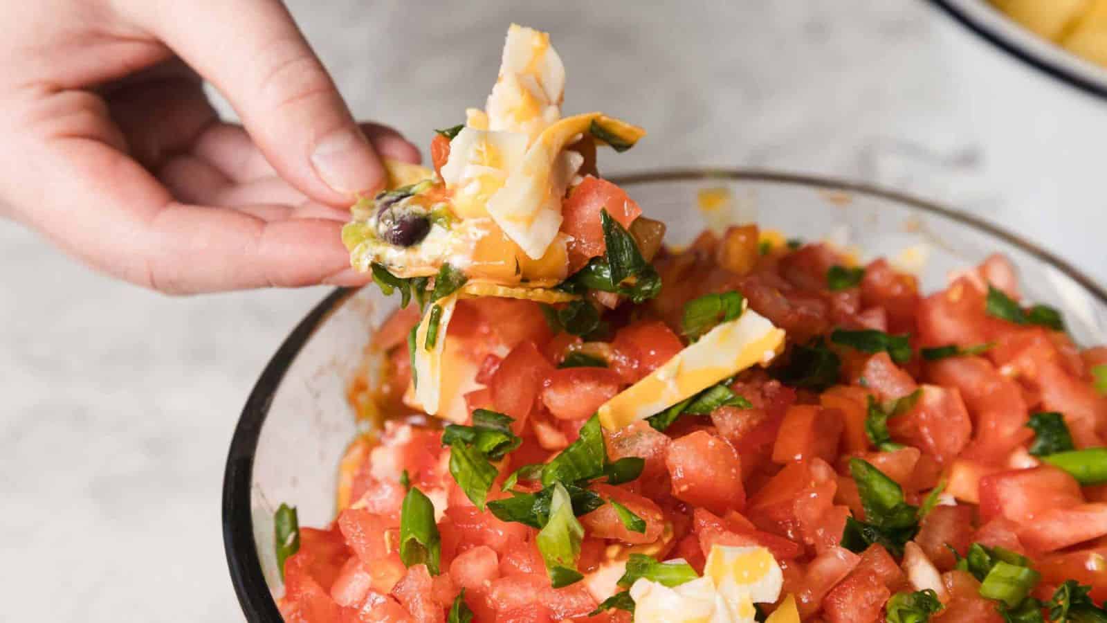 A person holds a chip topped with diced tomatoes, green onions, cheese, and olives, over a bowl filled with the same ingredients. The background is a light-colored countertop.