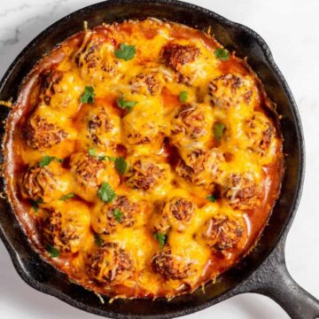 A cast iron skillet filled with baked meatballs covered in melted cheese and garnished with fresh herbs, placed on a white marble surface.