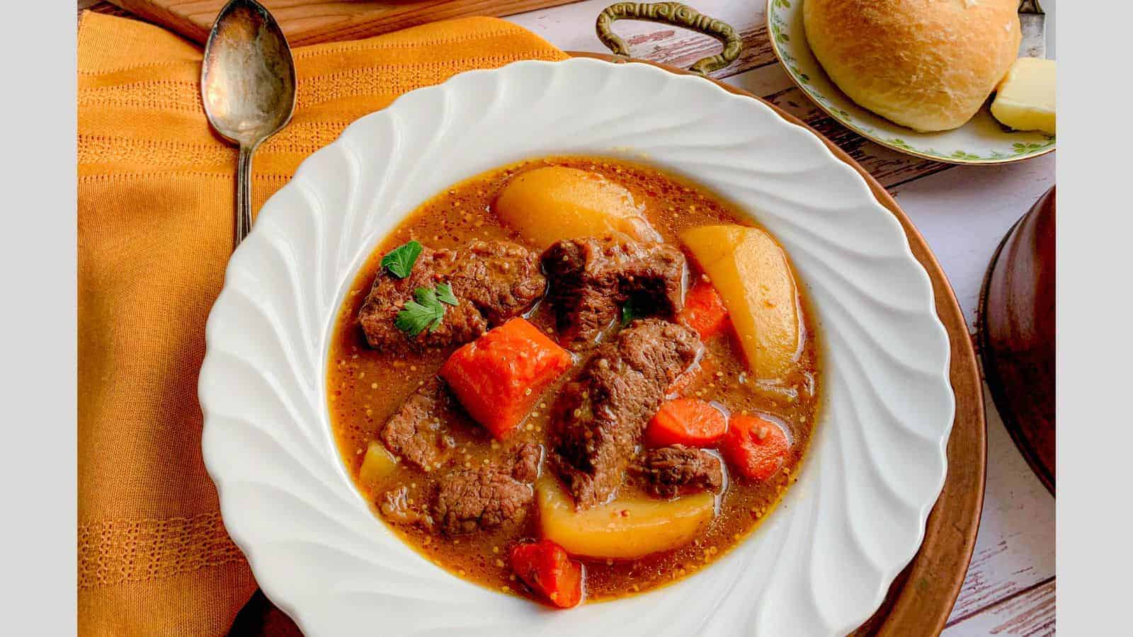 A white bowl brimming with beef stew—tender chunks of beef, potatoes, and carrots—garnished with parsley. Placed on a wooden surface beside an orange cloth, a spoon, and a bread roll; it brings back sweet memories of Grandma's cherished recipes.