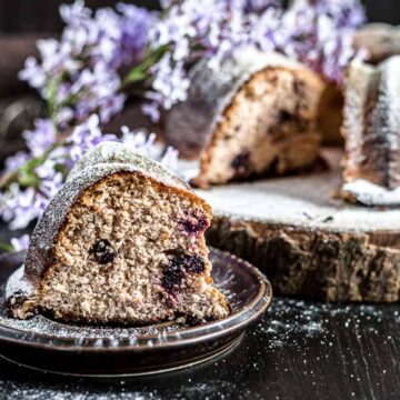 A slice of heavenly blueberry bundt cake sits on a small plate, tempting any sweet tooth. The rest of the cake rests majestically on a wooden board, lightly dusted with powdered sugar. Purple flowers decorate the dark surface, adding an elegant touch to this delightful scene of cakes.