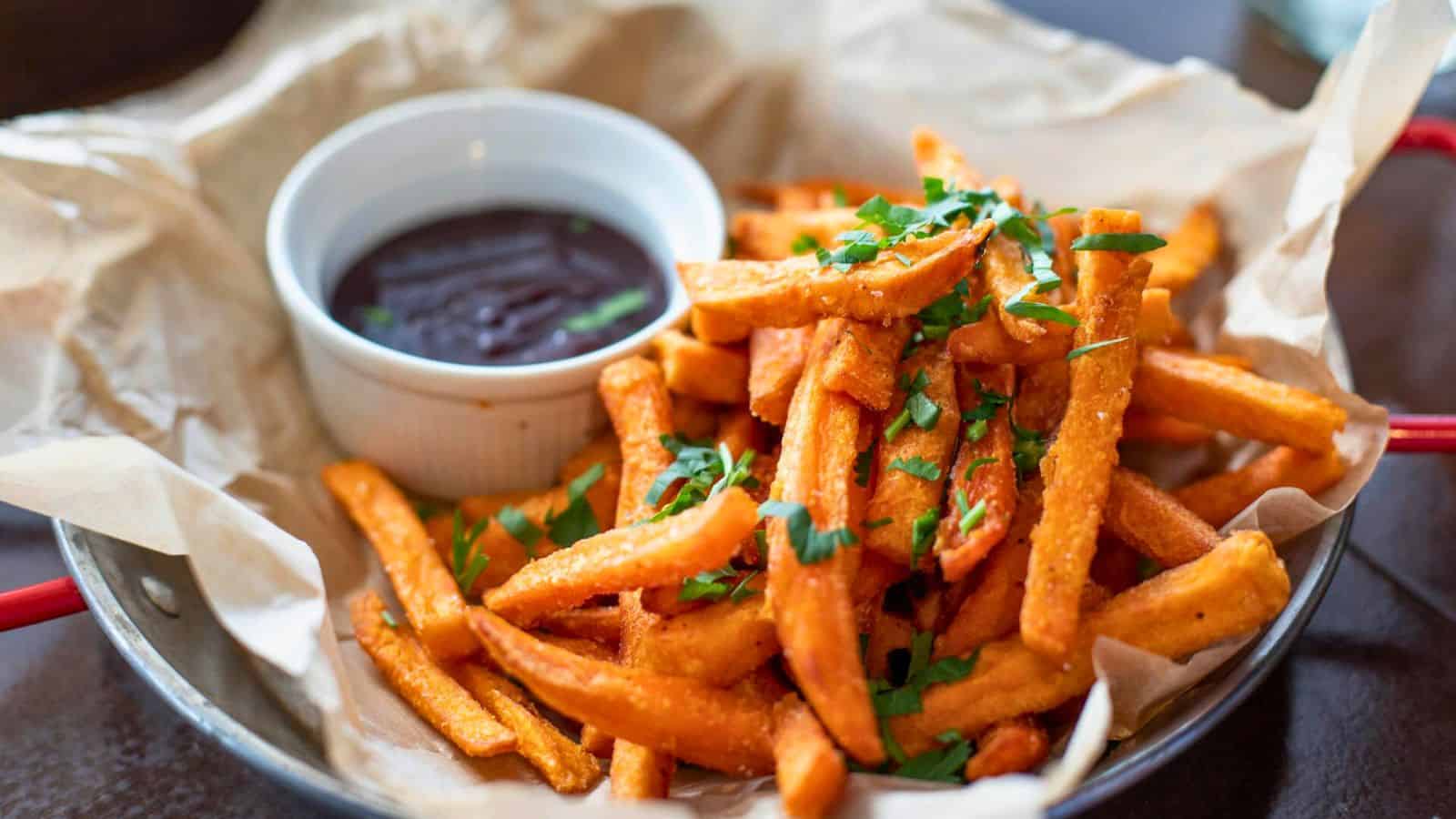 A basket of sweet potato fries garnished with chopped parsley, served on parchment paper. Accompanied by a small white ramekin filled with a dark dipping sauce.