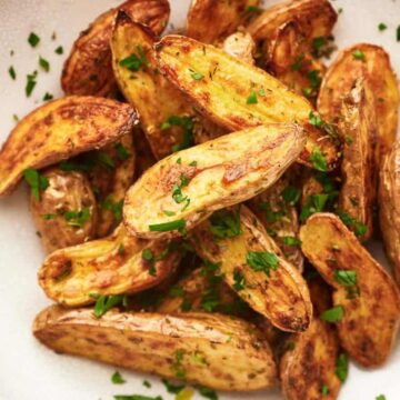 A white bowl filled with roasted potato wedges garnished with chopped parsley. The potatoes are golden-brown and neatly arranged, with visible seasoning and a hint of crisp texture. The bowl is set on a light-colored wooden surface.