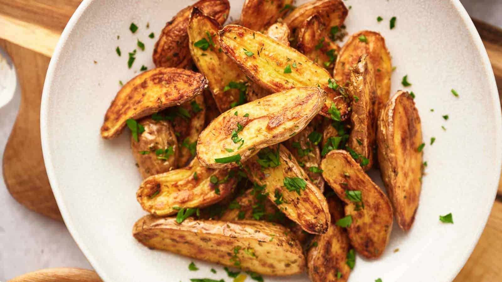 A white bowl filled with roasted potato wedges garnished with chopped parsley. The potatoes are golden-brown and neatly arranged, with visible seasoning and a hint of crisp texture. The bowl is set on a light-colored wooden surface.